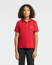 A young person with curly hair stands against a plain background, wearing the KIDS PIERCE PIQUE POLO - B0K201E200 from Psycho Bunny. This stylish red polo, crafted from rich Pima cotton, features an embroidered chest Bunny emblem and is perfectly paired with sleek black pants.