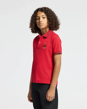 A young person with curly hair is wearing a vibrant red KIDS PIERCE PIQUE POLO (B0K201E200) by Psycho Bunny, standing against a plain background. The Pima cotton design boasts an embroidered Bunny on the chest, offering a touch of playful elegance to their look.