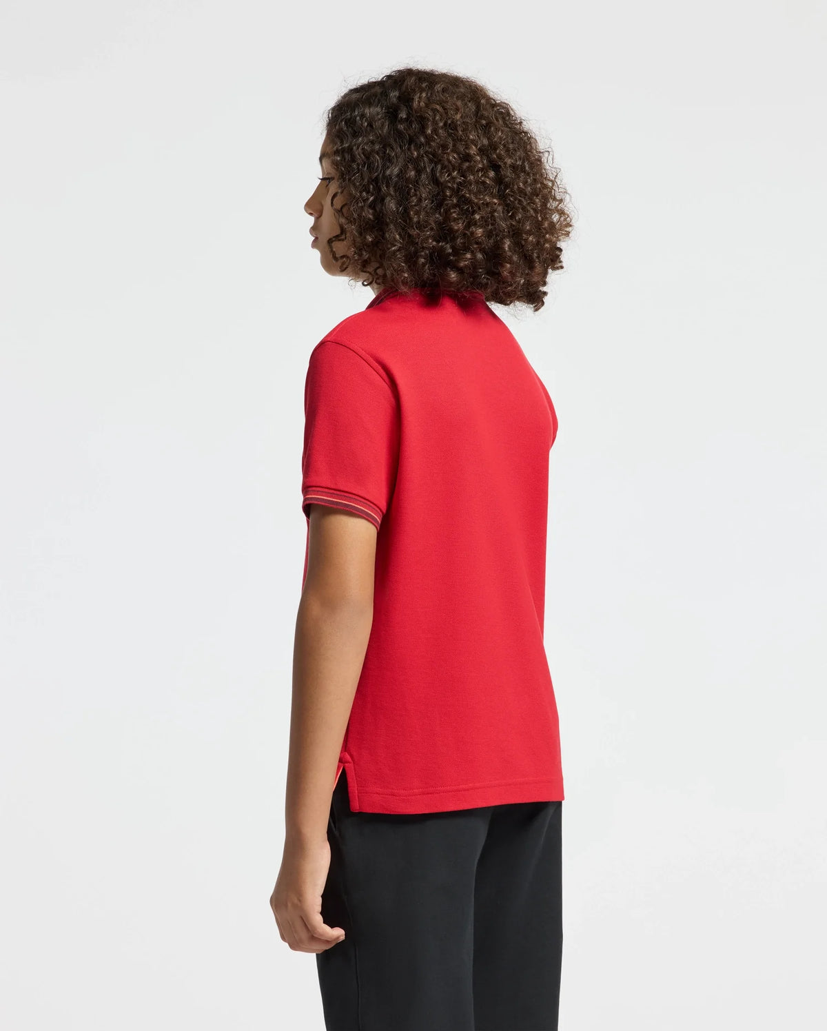 A person with curly hair is dressed in a red KIDS PIERCE PIQUE POLO - B0K201E200 by Psycho Bunny, showcasing an embroidered chest Bunny. They pair it with black pants and stand gracefully, gazing to the side against a plain background.