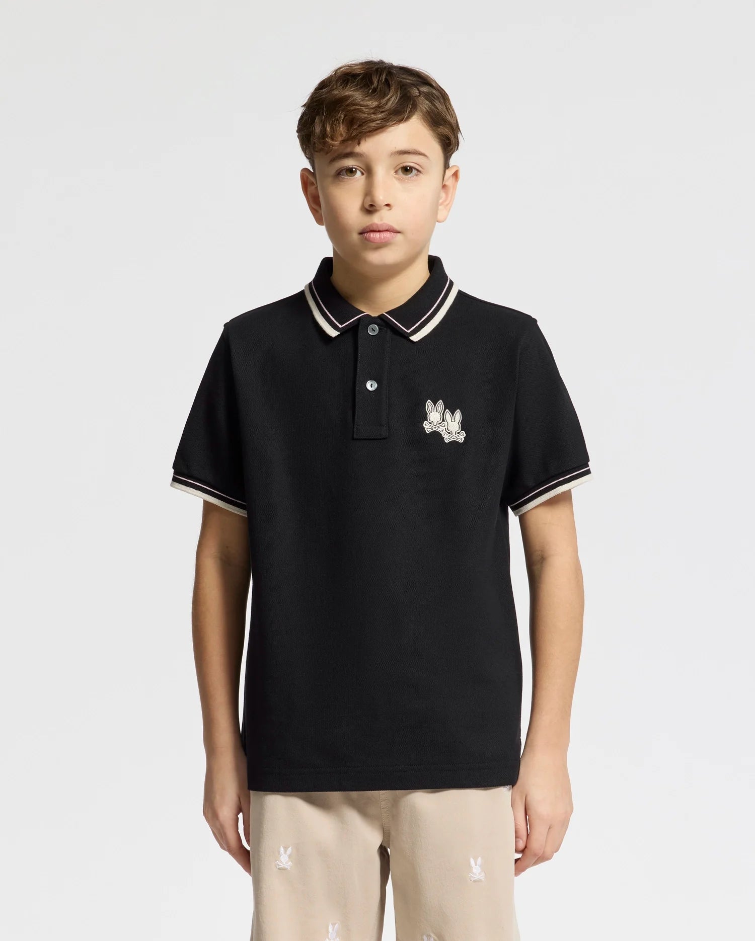 A young person models the black Psycho Bunny KIDS ELLIOT PIQUE POLO (B0K505F200), featuring white trim and an embroidered chest logo, against a plain white background. They have short brown hair and are looking directly at the camera, highlighting the polo's refined details.