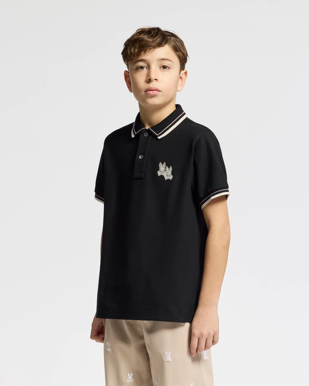 A young boy stands against a plain white background, wearing the Psycho Bunny KIDS ELLIOT PIQUE POLO made of soft Pima cotton with an embroidered logo on the chest, paired with light-colored pants that boast elevated finishes and a subtle pattern.