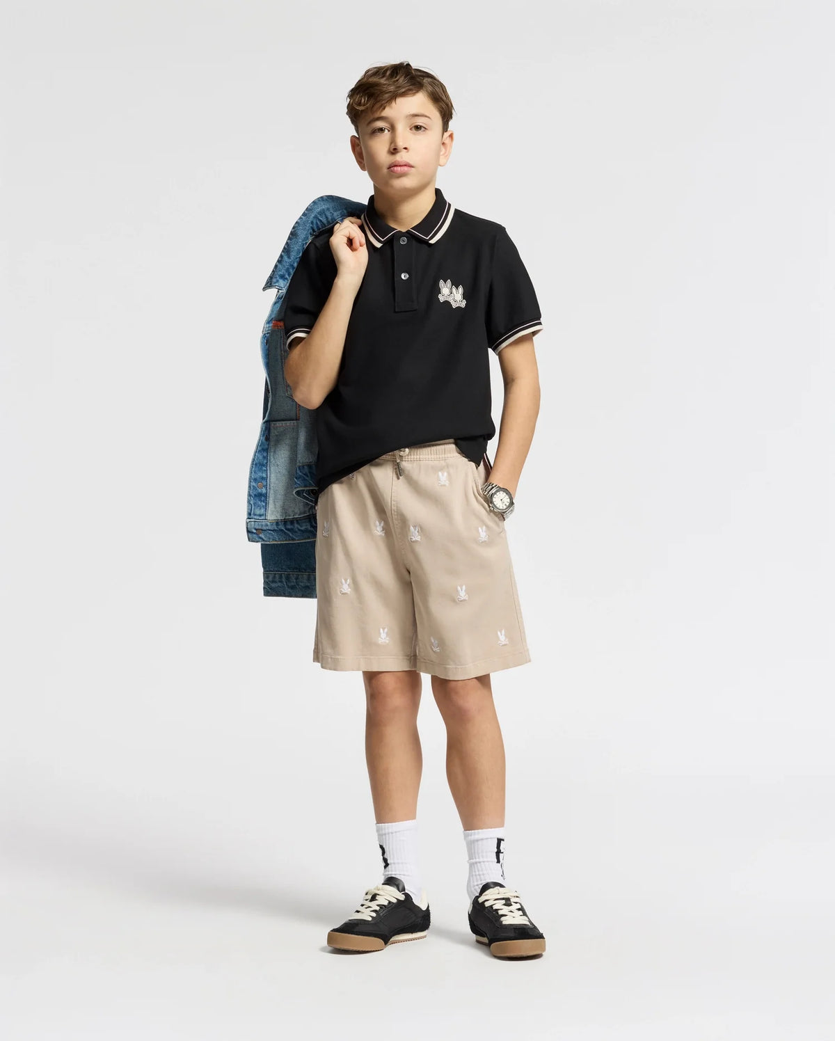 A young boy exudes style against a plain backdrop, with a denim jacket over his shoulder. He wears the KIDS ELLIOT PIQUE POLO from Psycho Bunny, beige patterned shorts, white socks, and black sneakers. A watch gleams on his wrist as he casually slips his left hand into his pocket.