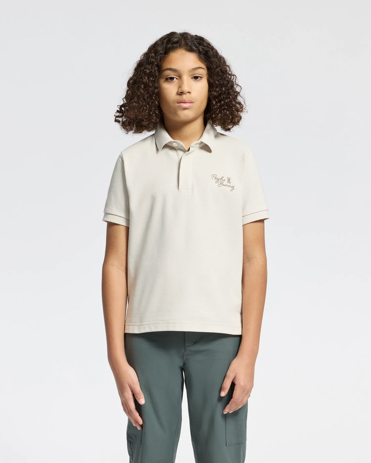 A person with curly hair stands facing forward against a plain background, wearing a Psycho Bunny KIDS DESERT PIQUE POLO in soft Pima cotton. The shirt has chest text and pairs elegantly with dark green pants. Their arms rest relaxed by their sides, exuding timeless style.