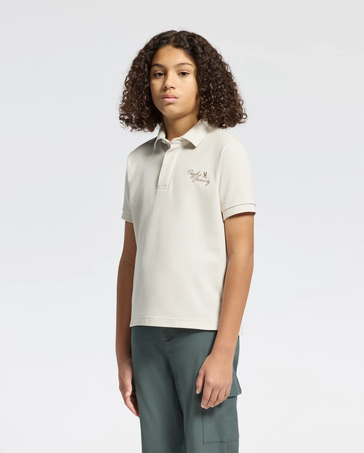 A young person with curly hair wears the KIDS DESERT PIQUE POLO by Psycho Bunny and dark green pants against a plain white background. Their hands rest at their sides while they exude timeless style and gaze confidently at the camera.