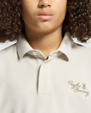 A person with curly hair is wearing the KIDS DESERT PIQUE POLO (B0K506F200) by Psycho Bunny in light beige. Made from Pima cotton, it showcases timeless style with a neatly folded collar and "Psycho Bunny" embroidered on the chest.