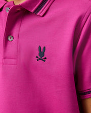 Close-up of a magenta KIDS RIVIERA PIQUE POLO SHIRT by Psycho Bunny, made from Pima cotton piqué, showcasing a black embroidered Bunny head and crossbones on the chest. The shirt features a buttoned collar and striped detailing.