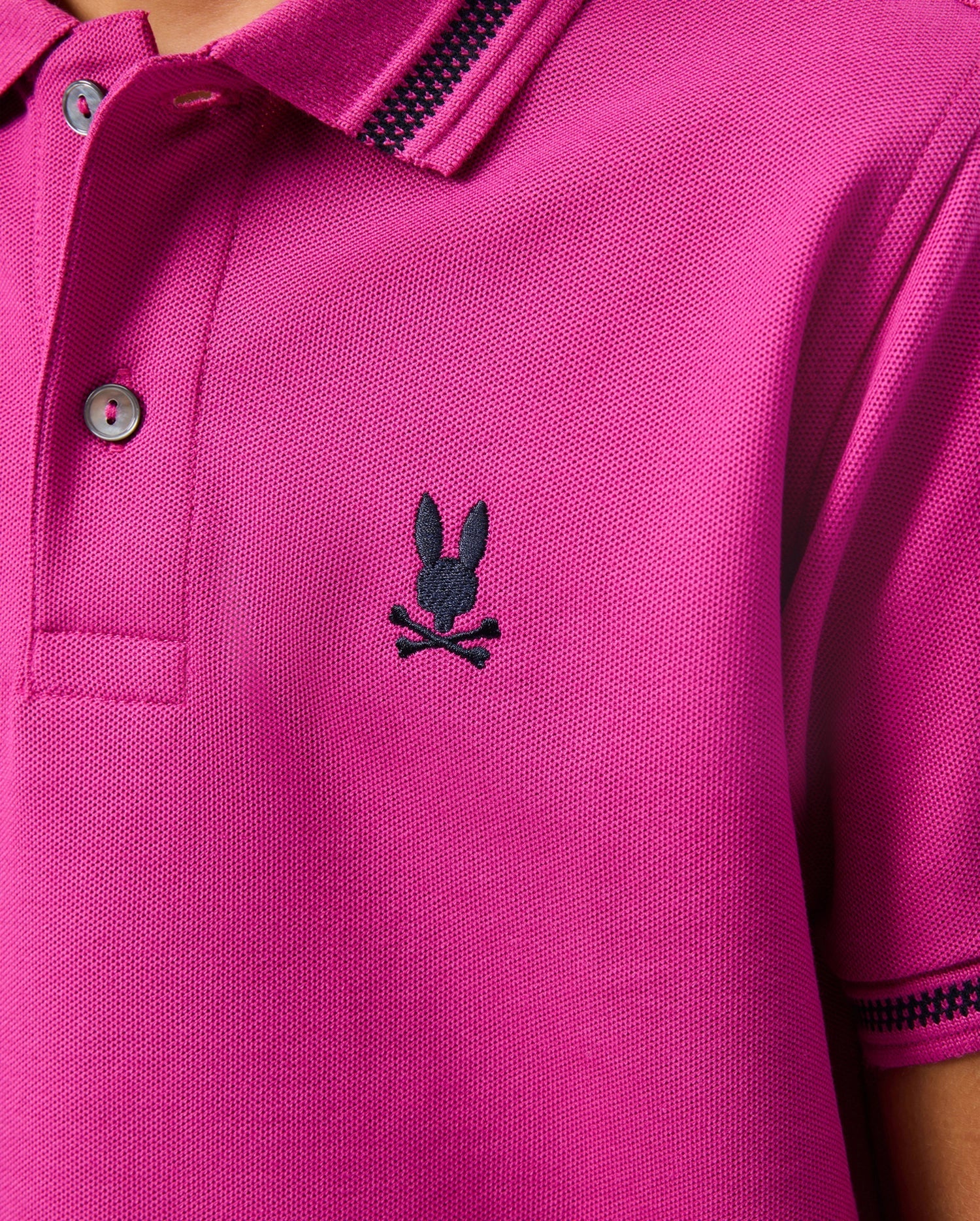 Close-up of a magenta KIDS RIVIERA PIQUE POLO SHIRT by Psycho Bunny, made from Pima cotton piqué, showcasing a black embroidered Bunny head and crossbones on the chest. The shirt features a buttoned collar and striped detailing.