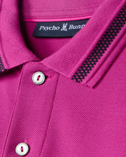 Here's a close-up of a vivid pink Kids Riviera Pique Polo Shirt (B0K640C200) by Psycho Bunny. It's made from Pima cotton piqué and features black and white striped details on the collar, buttons, and an embroidered Bunny logo on its small brand label.