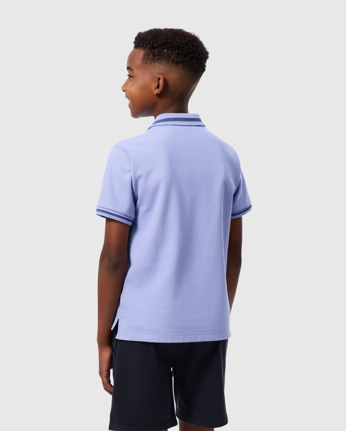 A boy, shown from behind, is wearing a light purple KIDS RIVIERA PIQUE POLO SHIRT - B0K640C200 by Psycho Bunny. The shirt features dark trim and is made from Pima cotton piqué, perfectly offsetting his short, curly hair against a plain gray background. He pairs the polo shirt with dark shorts.