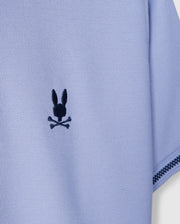Close-up of the sleeve of a KIDS RIVIERA PIQUE POLO SHIRT - B0K640C200 by Psycho Bunny, crafted from light blue textured fabric. It features a playful twist on the classic skull and crossbones with an embroidered black emblem of rabbit ears above crossed bones, echoing the whimsical charm of an embroidered chest Bunny.