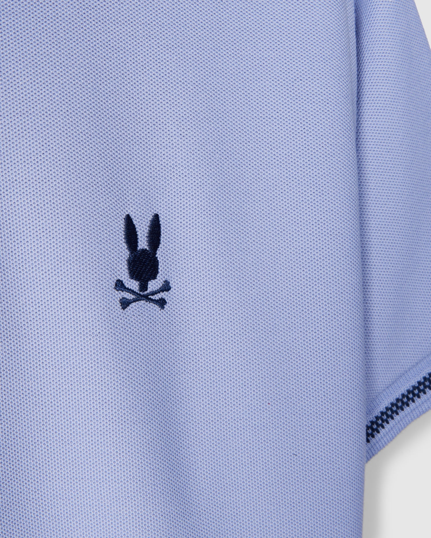 Close-up of the sleeve of a KIDS RIVIERA PIQUE POLO SHIRT - B0K640C200 by Psycho Bunny, crafted from light blue textured fabric. It features a playful twist on the classic skull and crossbones with an embroidered black emblem of rabbit ears above crossed bones, echoing the whimsical charm of an embroidered chest Bunny.