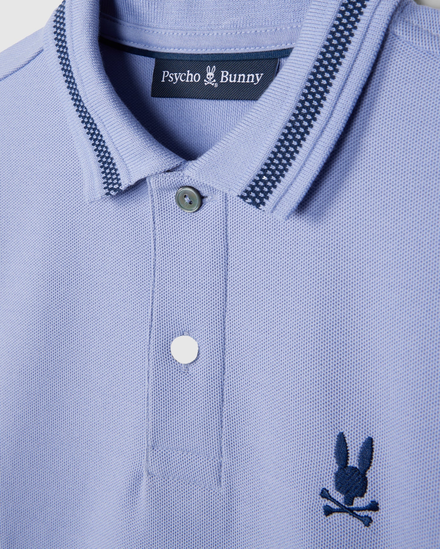 A close-up of the KIDS RIVIERA PIQUE POLO SHIRT - B0K640C200 by Psycho Bunny in light blue Pima cotton piqué showcases a dark blue embroidered chest Bunny logo with a bunny and crossbones. The collar features a striped pattern, and the shirt comes with two buttons.