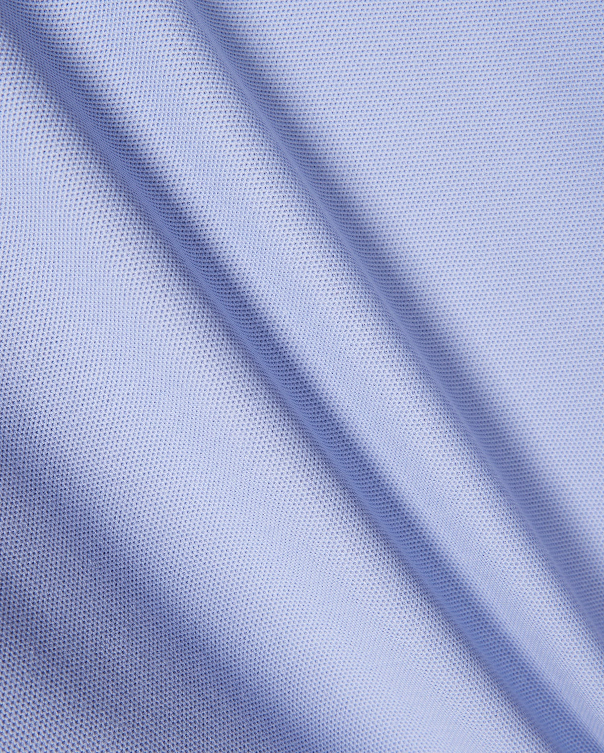 Close-up of light blue textured fabric with a fine mesh pattern, similar to Pima cotton piqué. The material shows gentle diagonal folds, indicative of its soft drape and smooth surface, echoing the refined elegance characteristic of the Psycho Bunny KIDS RIVIERA PIQUE POLO SHIRT - B0K640C200.