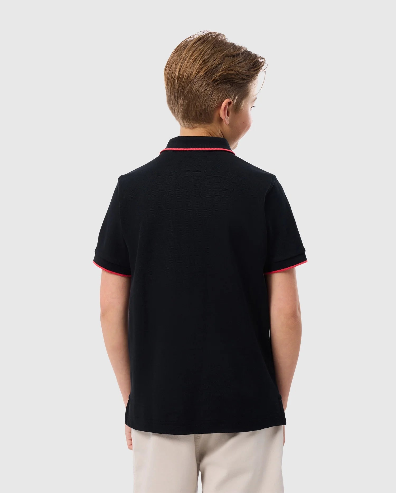 A young boy stands with his back to the camera, wearing a black KIDS KAYDEN PIQUE POLO - B0K675C200 from Psycho Bunny, featuring red trim on the collar and sleeves. Made from Pima cotton piqué, it complements his light brown hair and beige pants against a plain white background.