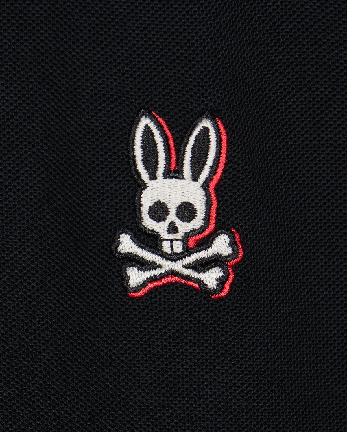 A logo featuring a white skull with rabbit ears and two crossed bones beneath it, embroidered on a black Pima cotton piqué fabric background. The skull and bones have red outline accents, adding striking detail to the bold contrasting stripes of the Psycho Bunny Kids Kayden Pique Polo (B0K675C200).
