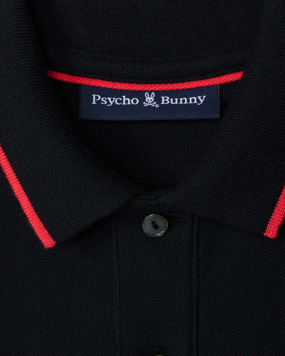 Close-up of the Kids Kayden Pique Polo (B0K675C200) by Psycho Bunny in black, featuring bold contrasting stripes of red trim on the collar. A label on the inside reads "Psycho Bunny," with a small bunny logo between the words. Made from Pima cotton piqué, this shirt has a buttoned collar with one button visible.