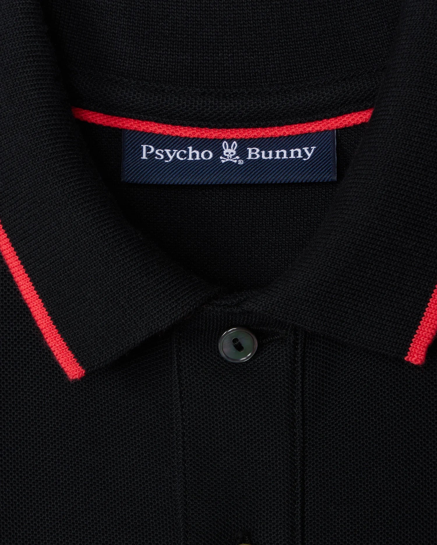 Close-up of the Kids Kayden Pique Polo (B0K675C200) by Psycho Bunny in black, featuring bold contrasting stripes of red trim on the collar. A label on the inside reads "Psycho Bunny," with a small bunny logo between the words. Made from Pima cotton piqué, this shirt has a buttoned collar with one button visible.