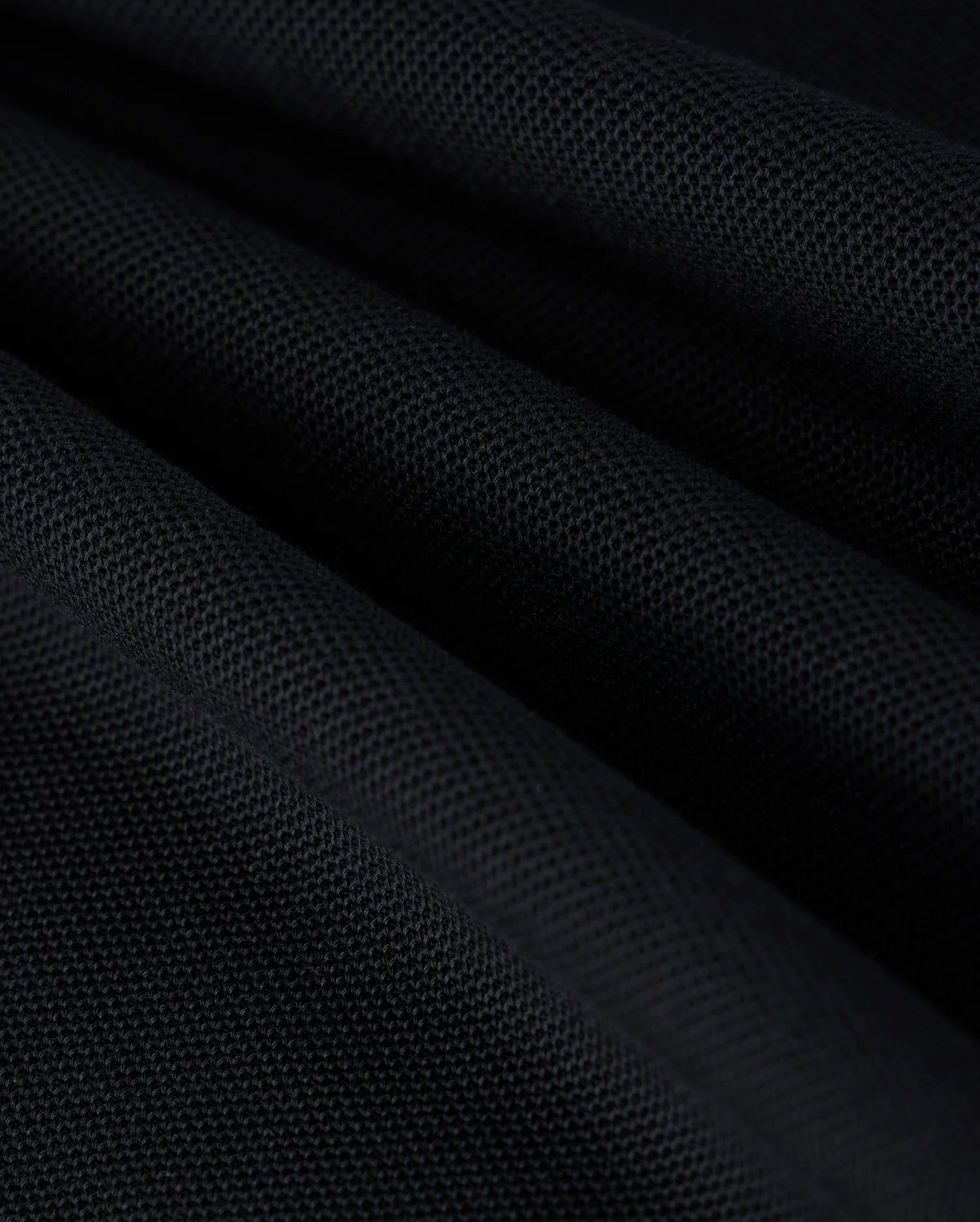 Close-up image of black Pima cotton piqué fabric with a textured surface, showcasing the intricate weave pattern. The fabric is arranged in folds, creating a play of light and shadow across its surface, reminiscent of the sophisticated elegance found in Psycho Bunny's KIDS KAYDEN PIQUE POLO - B0K675C200.