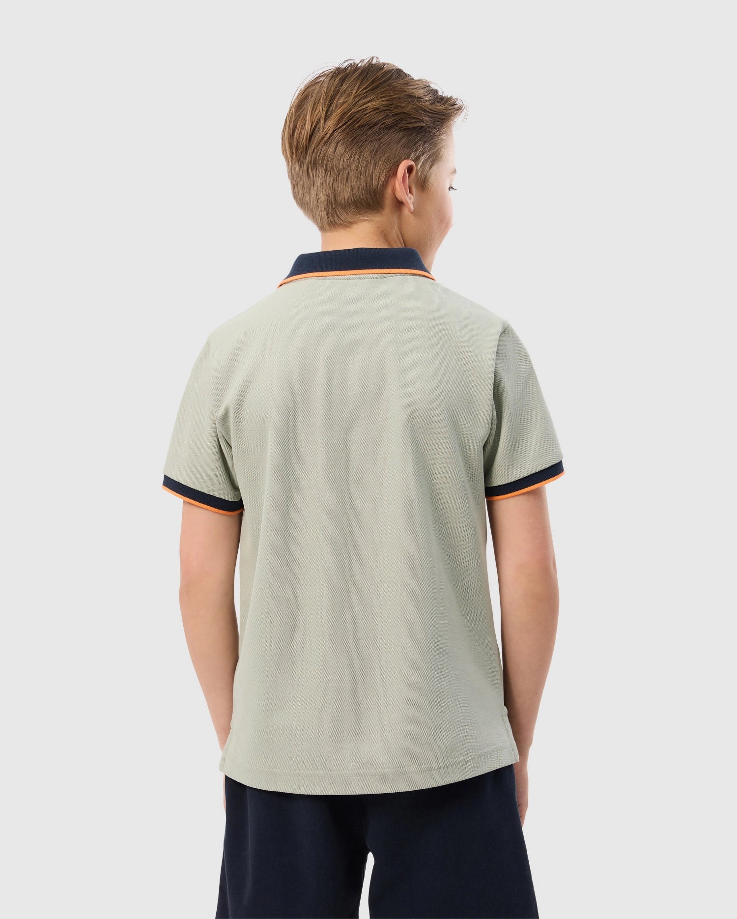 A young boy with short, light brown hair is seen from the back, wearing a Psycho Bunny KIDS KAYDEN PIQUE POLO - B0K675C200 in light gray with dark navy collar and sleeve trims, featuring contrasting stripes. The Pima cotton pique shirt pairs perfectly with his dark navy shorts as he stands against a plain, light gray background.