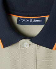 A close-up of the Kids Kayden Pique Polo - B0K675C200, featuring a khaki color with a black collar and orange trim, crafted from soft Pima cotton pique. The shirt displays the Psycho Bunny brand label inside the collar, showcasing the iconic bunny logo with crossbones below it. A single white button is visible beneath contrasting stripes on the placket.