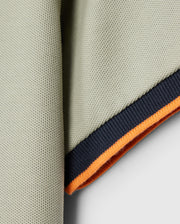 A close-up of the corner of the Psycho Bunny KIDS KAYDEN PIQUE POLO - B0K675C200, highlighting its Pima cotton pique fabric with a ribbed edge. The edge features contrasting stripes: a dark blue stripe followed by a bright orange band underneath, set against a neutral, light gray background.