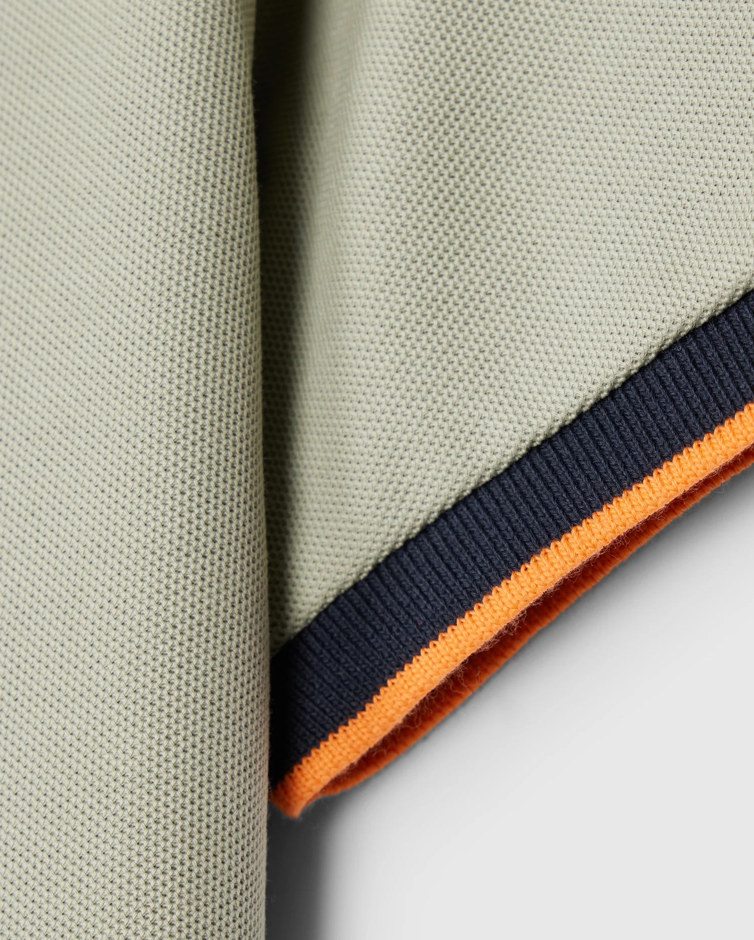 A close-up of the corner of the Psycho Bunny KIDS KAYDEN PIQUE POLO - B0K675C200, highlighting its Pima cotton pique fabric with a ribbed edge. The edge features contrasting stripes: a dark blue stripe followed by a bright orange band underneath, set against a neutral, light gray background.