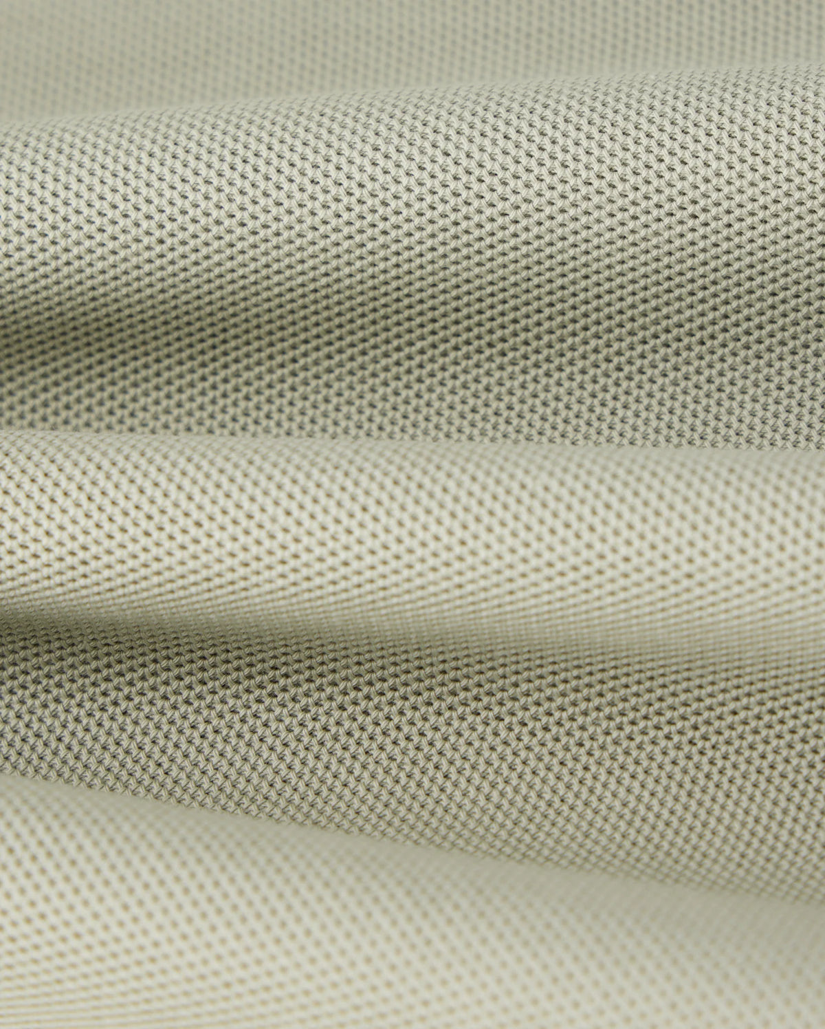 Close-up of a piece of light gray, finely textured Pima cotton pique fabric from the Psycho Bunny KIDS KAYDEN PIQUE POLO - B0K675C200, with a slightly rough surface. The fabric appears to be folded, creating gentle, undulating waves. The material's weave is visible, featuring a uniform pattern of small, evenly spaced dots.