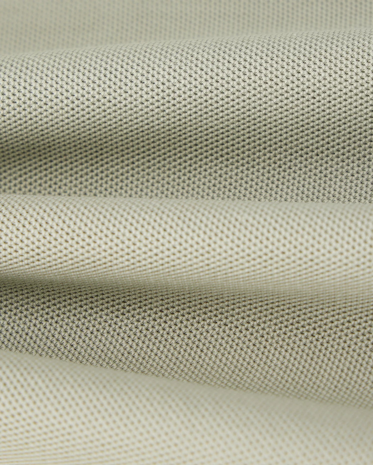 Close-up of a piece of light gray, finely textured Pima cotton pique fabric from the Psycho Bunny KIDS KAYDEN PIQUE POLO - B0K675C200, with a slightly rough surface. The fabric appears to be folded, creating gentle, undulating waves. The material's weave is visible, featuring a uniform pattern of small, evenly spaced dots.