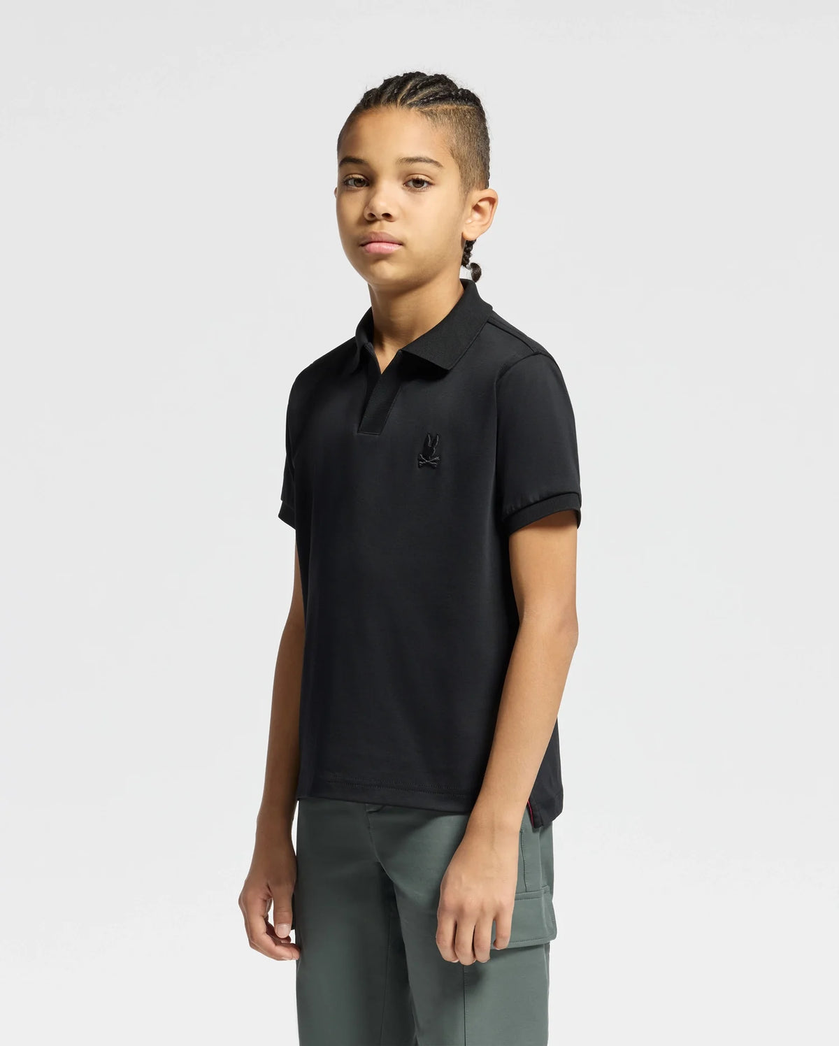 A young person with braided hair is wearing a black Psycho Bunny Kids Broadway Pima Interlock Johnny Collar Polo and green pants, standing against a plain white background.
