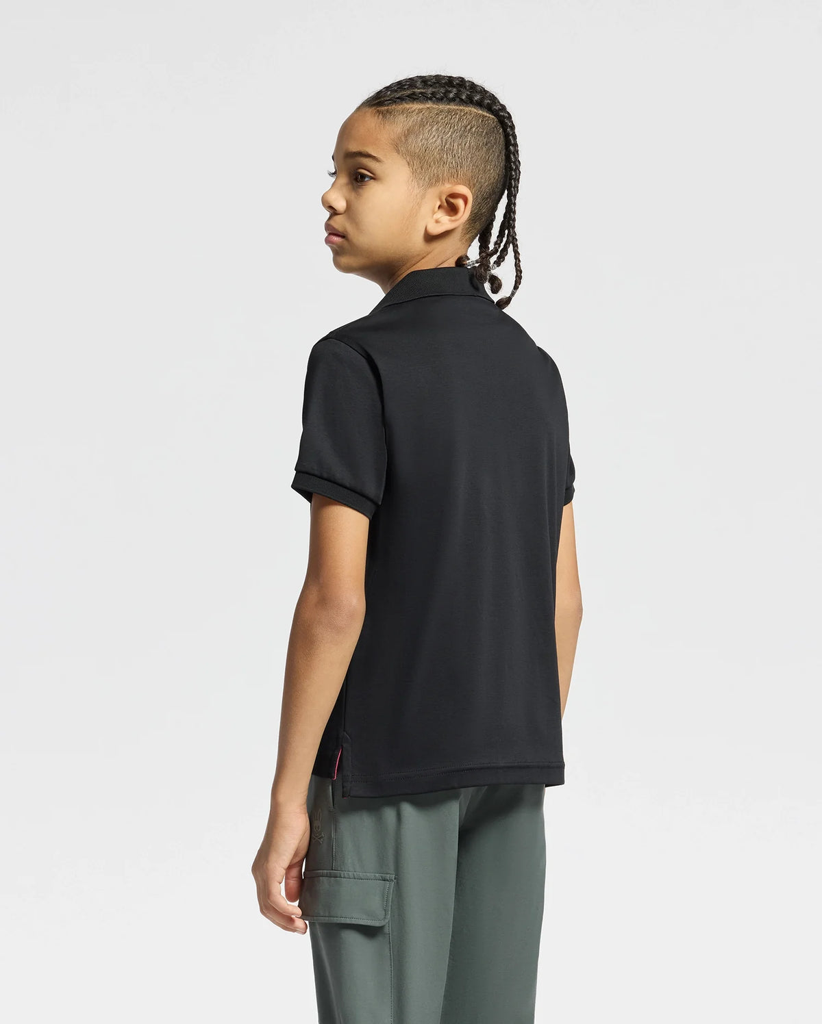 A young person with braids stands in a three-quarter view, facing away from the camera. They wear a Psycho Bunny KIDS BROADWAY PIMA INTERLOCK JOHNNY COLLAR POLO in black and gray pants with a side pocket, all against a plain white background.