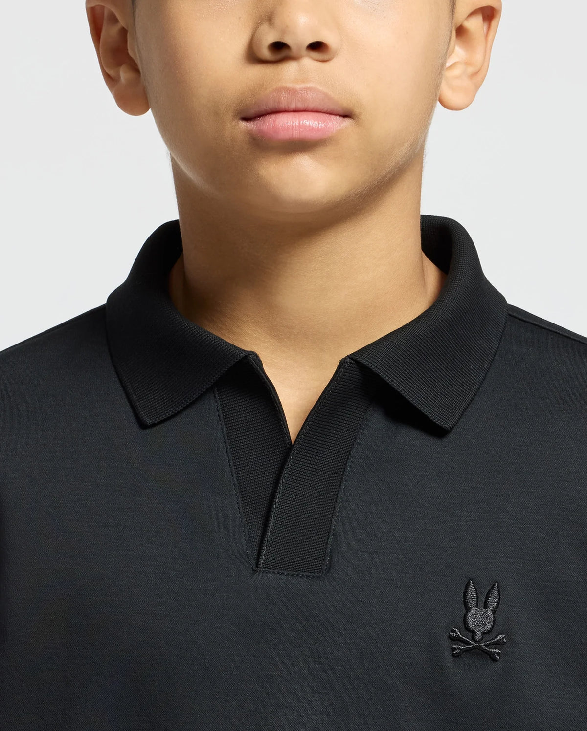 A person is wearing a black Psycho Bunny Kids Broadway Pima Interlock Johnny Collar Polo, showcasing a small embroidered rabbit with crossbones logo on the chest. The image highlights the sleek design of the upper torso and neck area.
