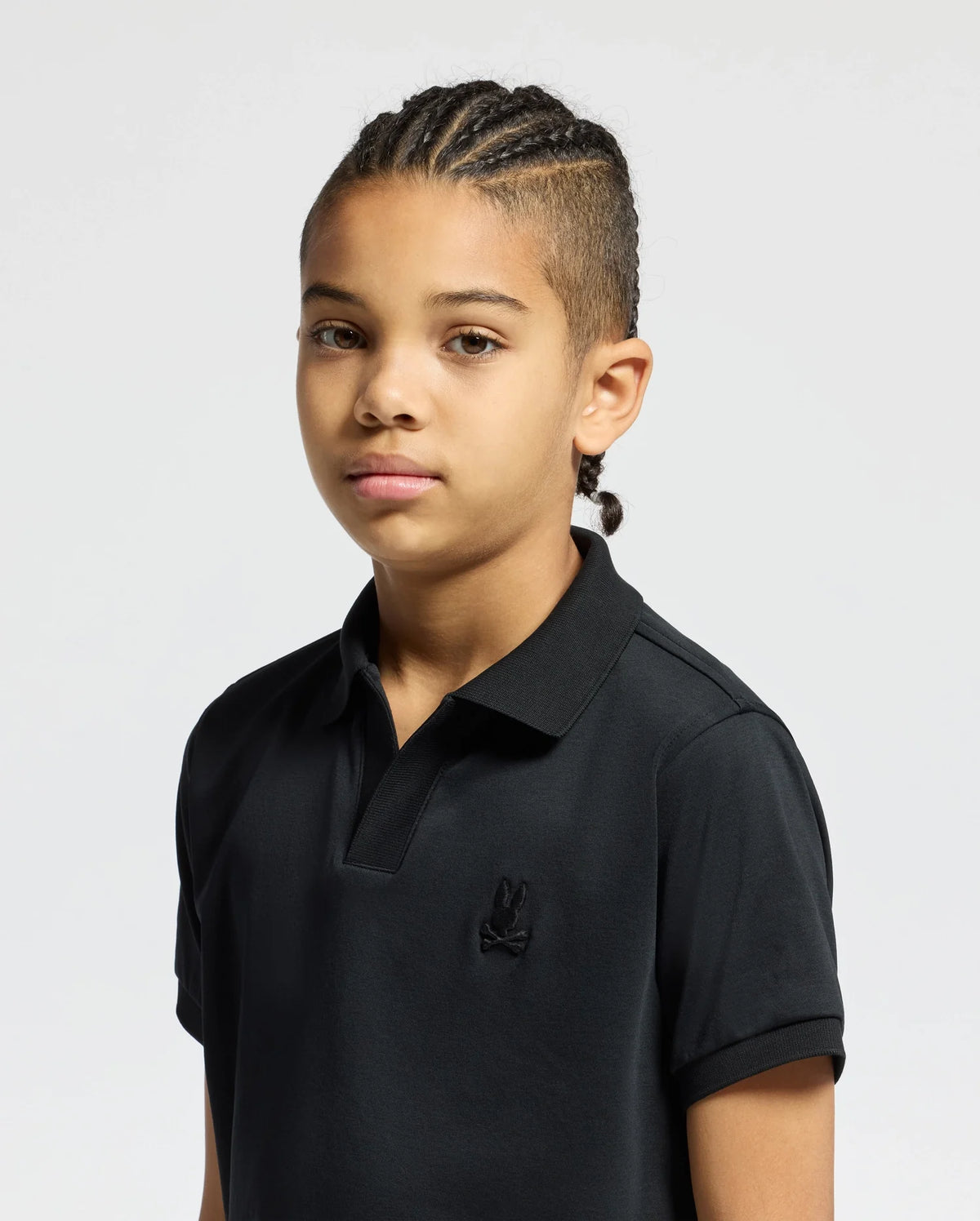 A young person with braided hair wears a black Psycho Bunny KIDS BROADWAY PIMA INTERLOCK JOHNNY COLLAR POLO (B0K891D200), standing against a plain background, gazing directly ahead.