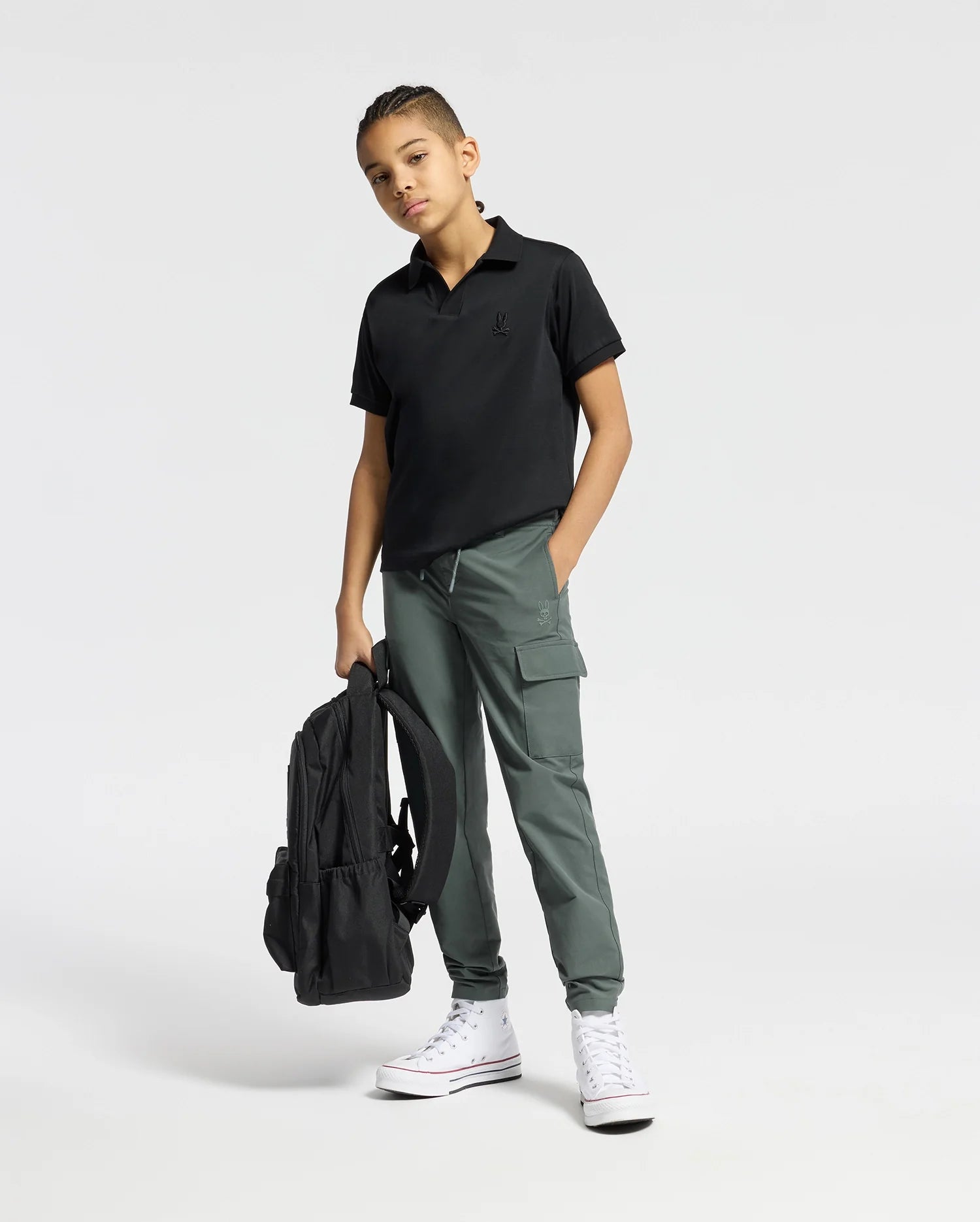 A young person stands against a plain background, wearing the stylish Psycho Bunny Kids Broadway Pima Interlock Johnny Collar Polo and green cargo pants. They hold a black backpack, sport white high-top sneakers, and have braided hair for an effortlessly cool look.