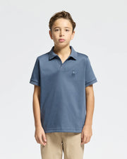 A young boy is posed against a plain background, dressed in Psycho Bunny's KIDS BROADWAY PIMA INTERLOCK JOHNNY COLLAR POLO (B0K891D200) in blue, highlighted by a small chest logo, and beige pants. The mercerized Pima cotton shirt enhances his simple yet stylish look.