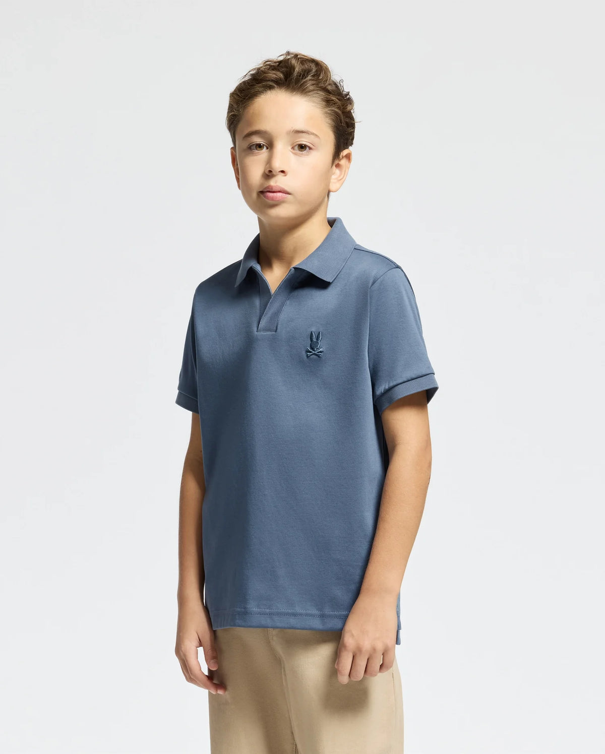 A young individual stands confidently against a plain background, wearing a Psycho Bunny KIDS BROADWAY PIMA INTERLOCK JOHNNY COLLAR POLO with a subtle logo on the left. They pair it with beige pants and short hair, gazing directly at the camera.