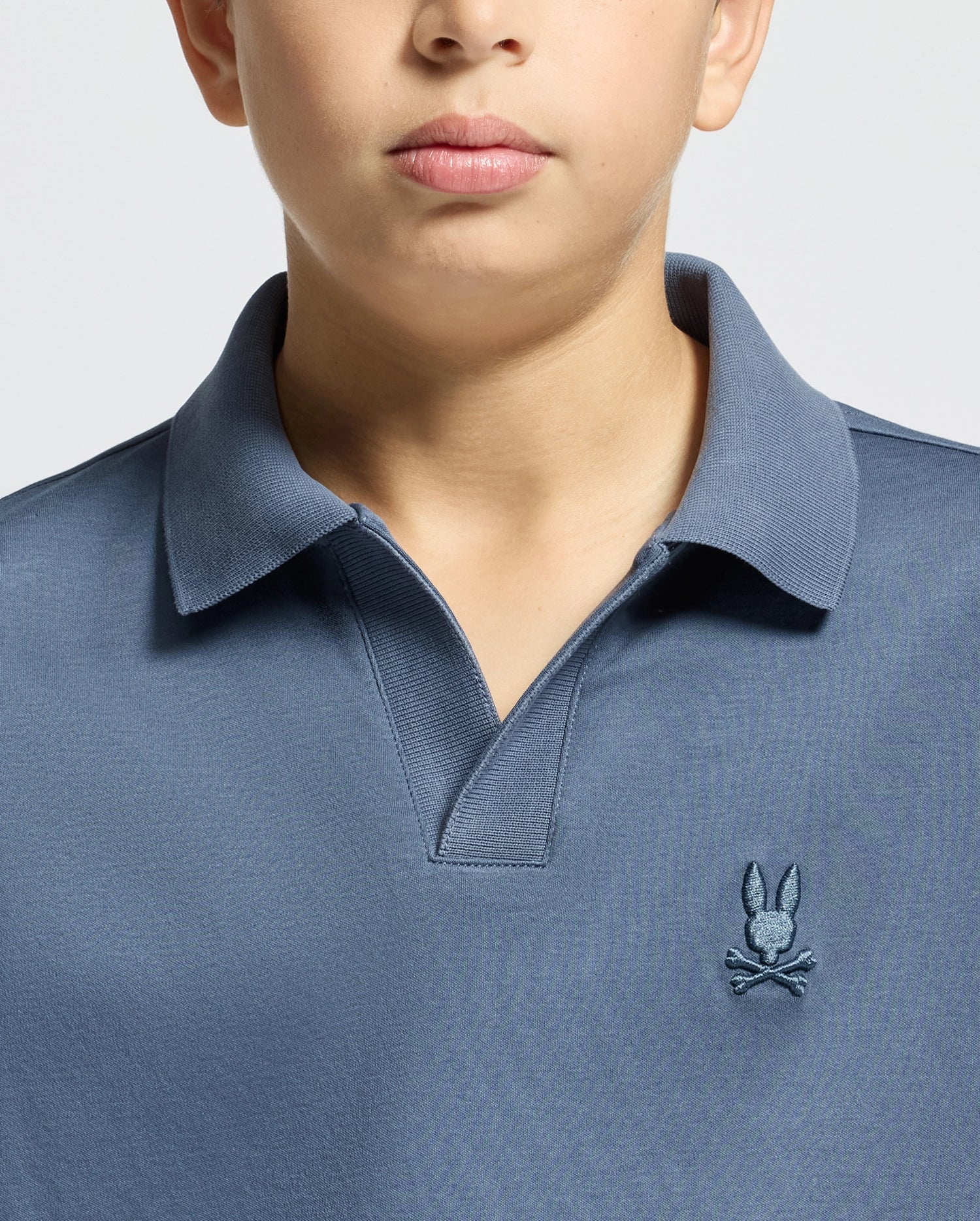 A child wearing a blue "KIDS BROADWAY PIMA INTERLOCK JOHNNY COLLAR POLO - B0K891D200" by Psycho Bunny, made from soft mercerized Pima cotton and featuring a small bunny and crossbones logo on the chest. The image highlights the upper torso and collar.