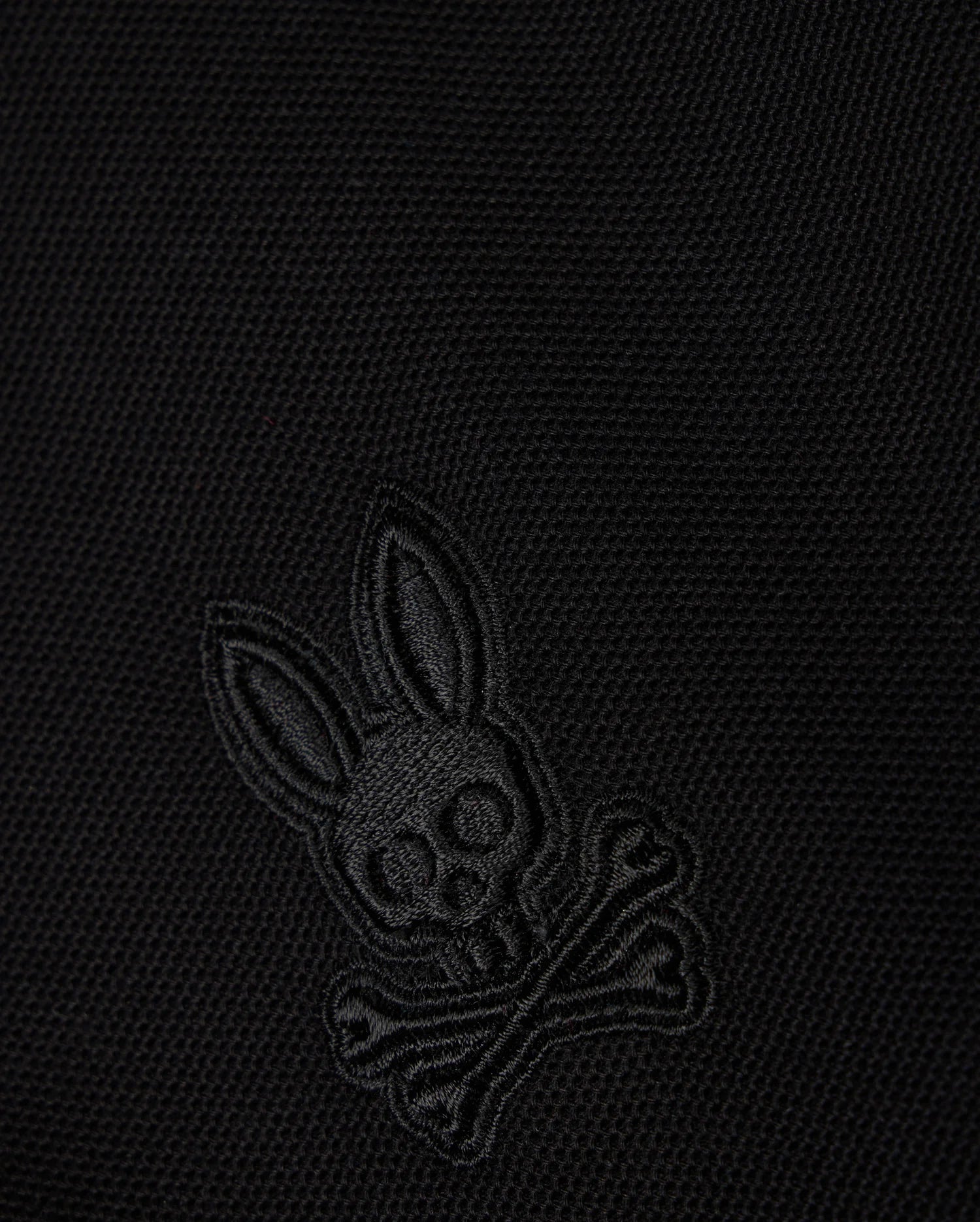 Close-up of a black Pima cotton piqué fabric with an embroidered Psycho Bunny logo on the KIDS MALTON PIQUE POLO SHIRT - B0K950D200: featuring a skull with elongated bunny ears above two crossed bones, all in a matching black color.