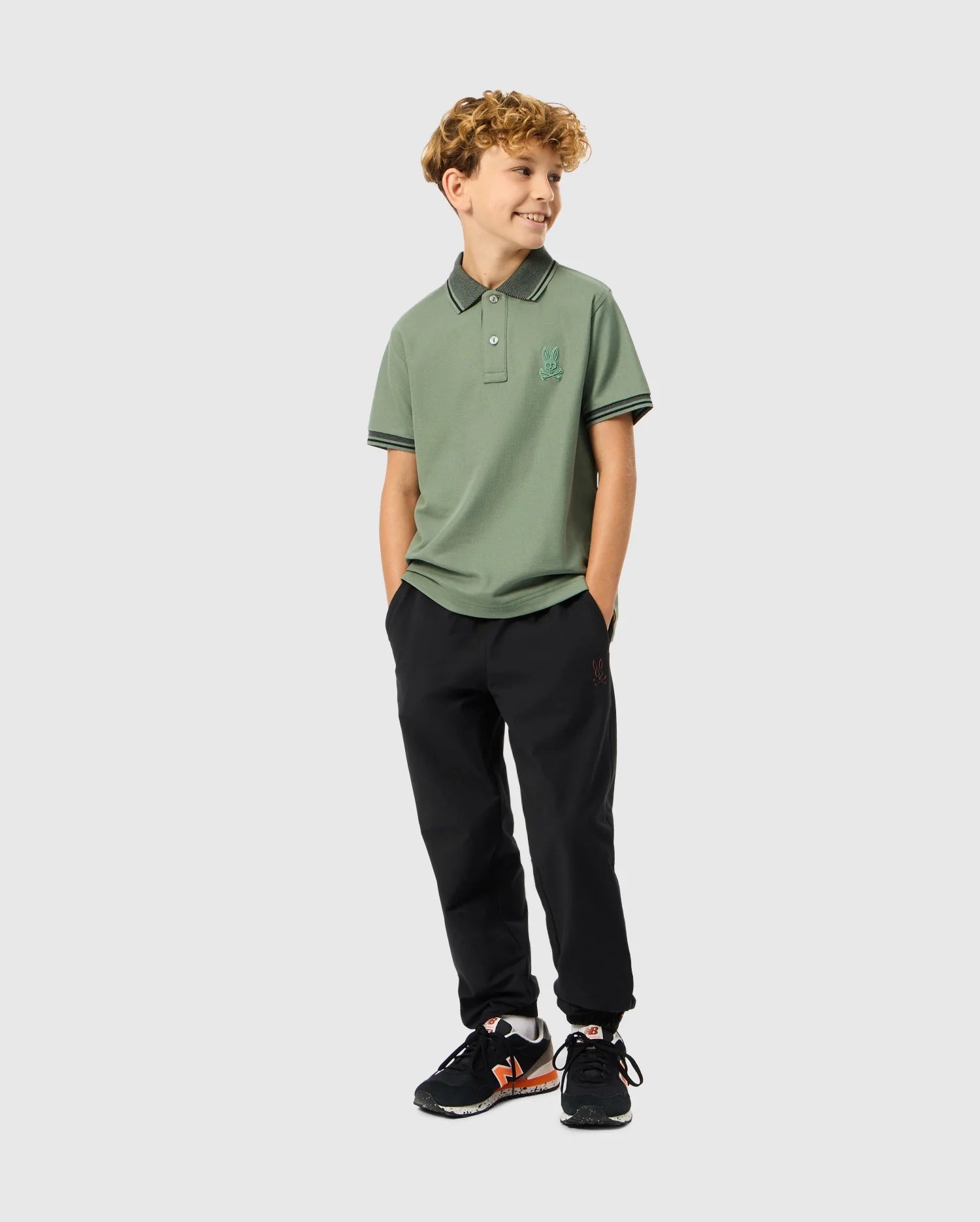 A young boy wearing a green Psycho Bunny KIDS MALTON PIQUE POLO SHIRT - B0K950D200, featuring diamond piqué-patterned cuffs, and black pants smiles while standing with his hands in his pockets. He has curly hair and is wearing black sneakers with orange accents against a plain white background.