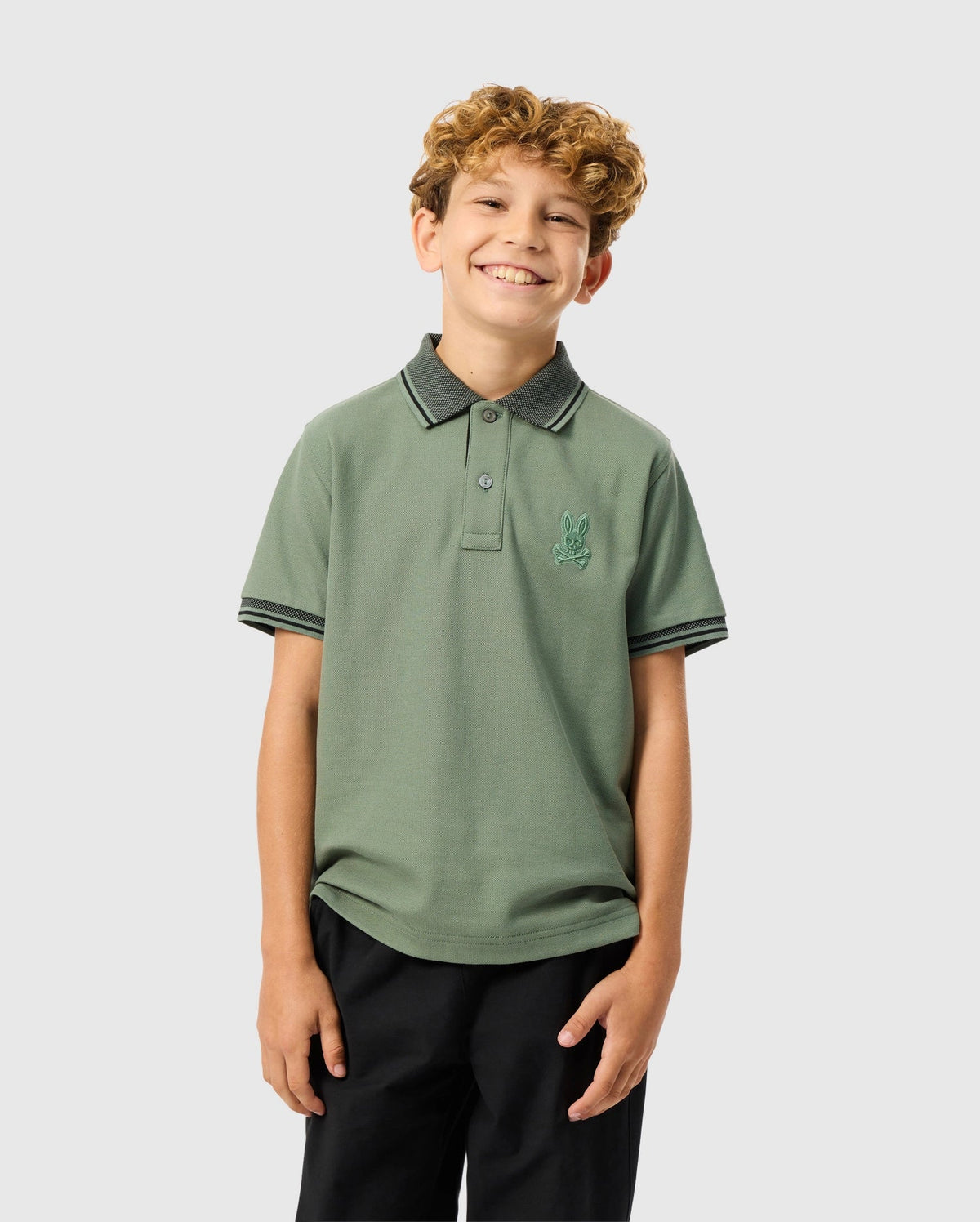A smiling young boy with curly hair wears a light green Psycho Bunny KIDS MALTON PIQUE POLO SHIRT - B0K950D200 made from pima cotton, showcasing diamond piqué-patterned cuffs and an embroidered logo on the left side. He pairs it with dark pants against a plain, light gray background.