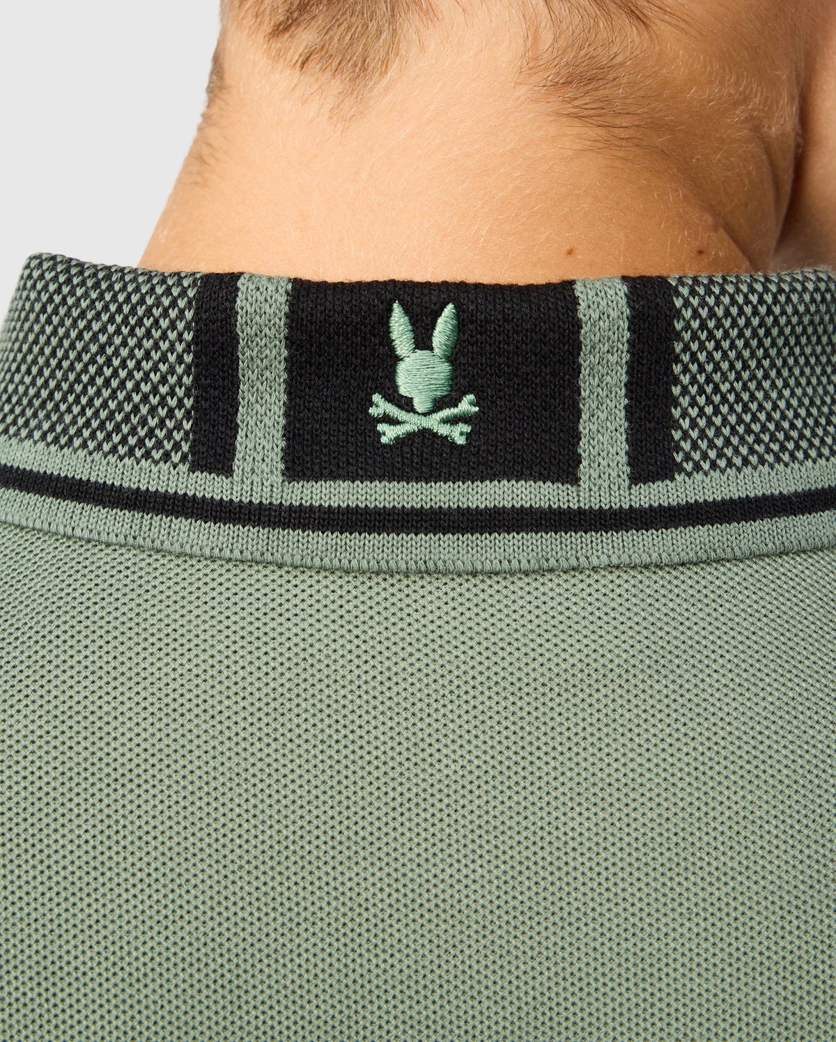 Close-up of the back of a neck wearing a green textured shirt crafted from soft pima cotton, featuring a black and green collar. The collar is adorned with a green embroidered bunny and crossbones logo, adding character to this Kids Malton Pique Polo Shirt by Psycho Bunny.