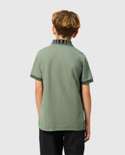 A young boy with light brown hair stands facing away from the camera, dressed in a green Psycho Bunny Kids Malton Pique Polo Shirt (B0K950D200) featuring diamond piqué-patterned cuffs and black trim, paired with black pants against a simple light gray backdrop.