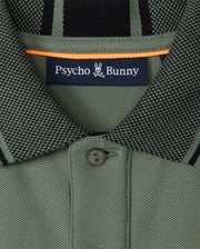 Close-up of a green collared shirt from the Psycho Bunny brand, made with soft pima cotton and featuring a small button. The label displays "Psycho Bunny," accompanied by the distinctive bunny skull logo. This KIDS MALTON PIQUE POLO SHIRT (B0K950D200) is adorned with black and orange accents and showcases diamond piqué-patterned cuffs for added style.