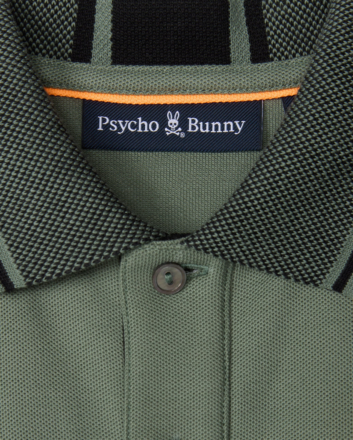 Close-up of a green collared shirt from the Psycho Bunny brand, made with soft pima cotton and featuring a small button. The label displays "Psycho Bunny," accompanied by the distinctive bunny skull logo. This KIDS MALTON PIQUE POLO SHIRT (B0K950D200) is adorned with black and orange accents and showcases diamond piqué-patterned cuffs for added style.