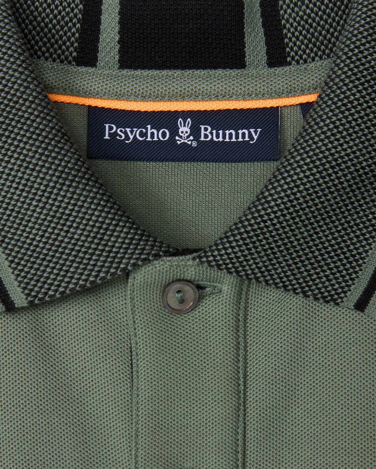 Close-up of a green collared shirt from the Psycho Bunny brand, made with soft pima cotton and featuring a small button. The label displays "Psycho Bunny," accompanied by the distinctive bunny skull logo. This KIDS MALTON PIQUE POLO SHIRT (B0K950D200) is adorned with black and orange accents and showcases diamond piqué-patterned cuffs for added style.