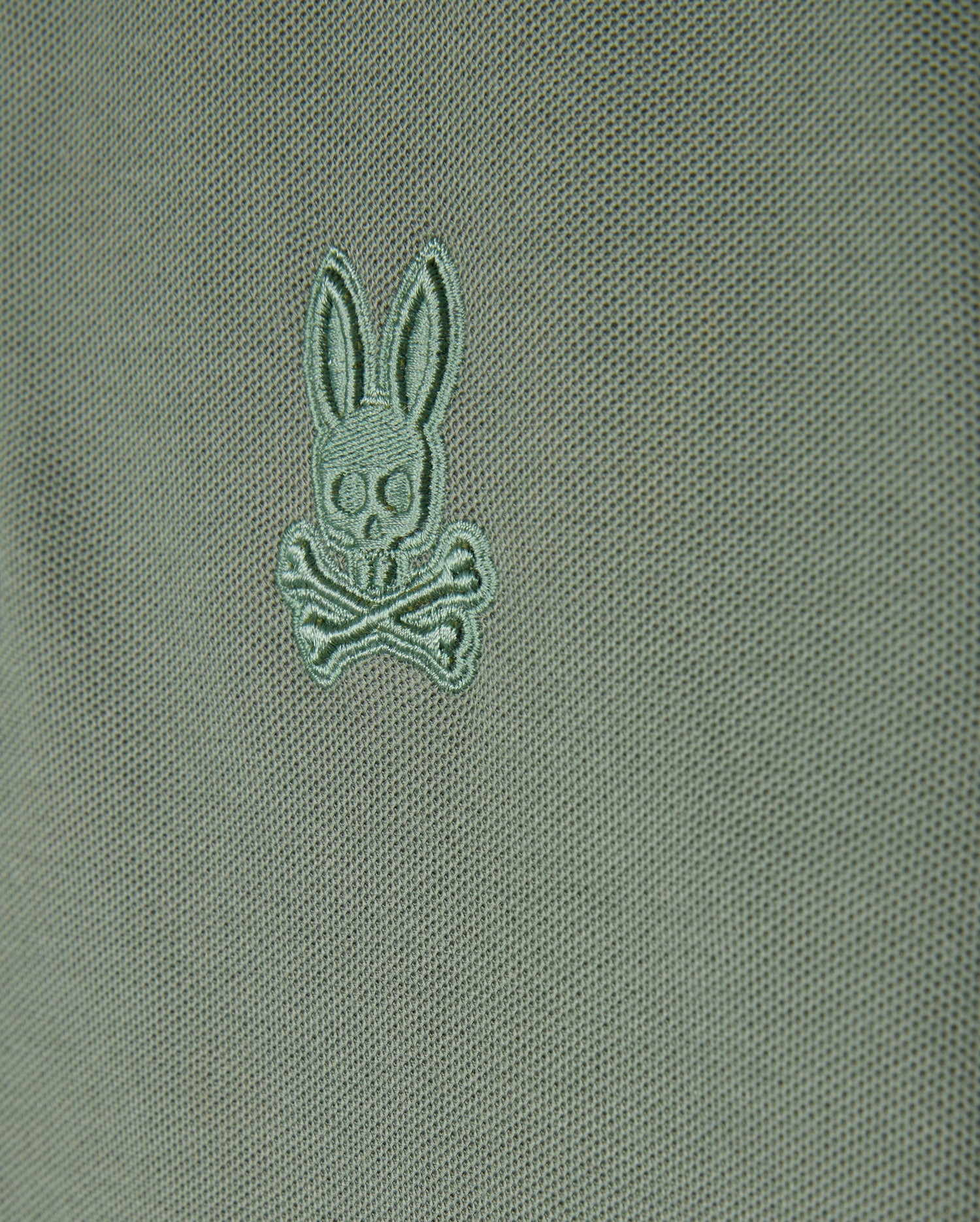 Close-up of a green Kids Malton Piqué Polo Shirt by Psycho Bunny, crafted from soft pima cotton. It showcases a small embroidered design featuring a stylized rabbit's head with crossed bones underneath. The shirt is enhanced with distinctive diamond piqué-patterned cuffs for an added touch of sophistication.