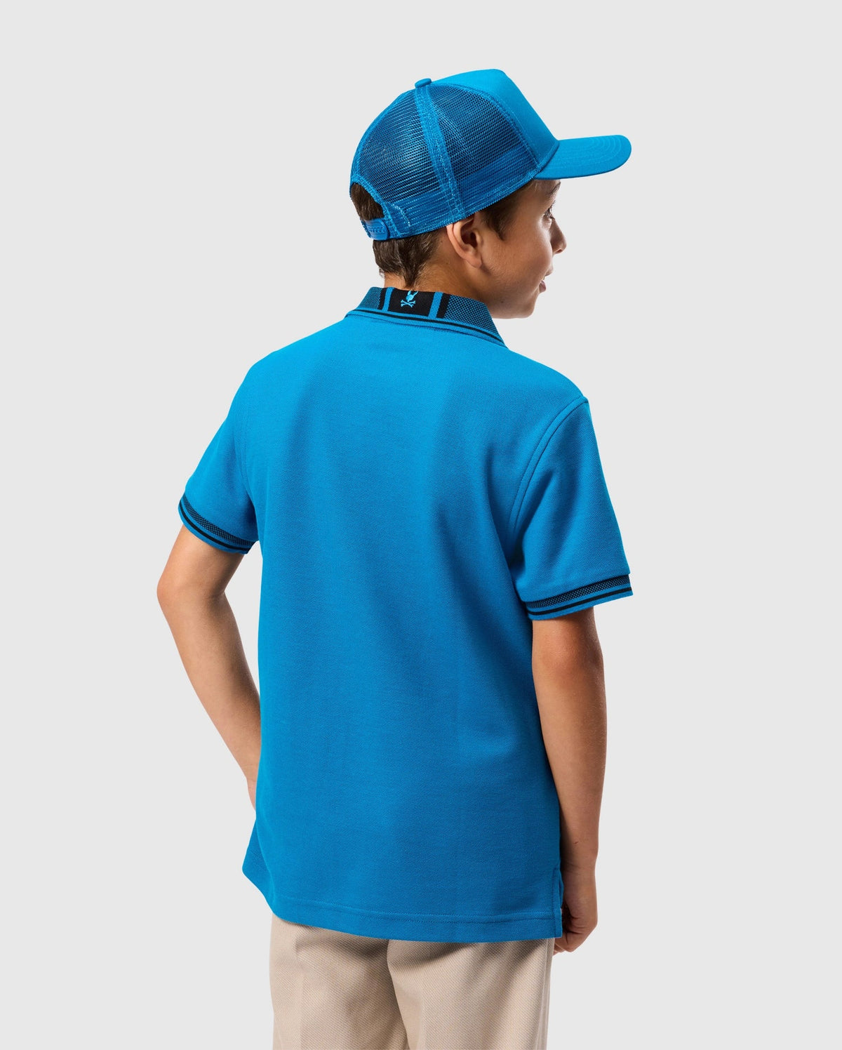 A boy is seen from behind wearing a Psycho Bunny KIDS MALTON PIQUE POLO SHIRT - B0K950D200, crafted from Pima cotton piqué, and featuring dark trim on the collar and sleeves along with mother-of-pearl buttons. He is also wearing a matching blue baseball cap, all set against a plain, light gray background.