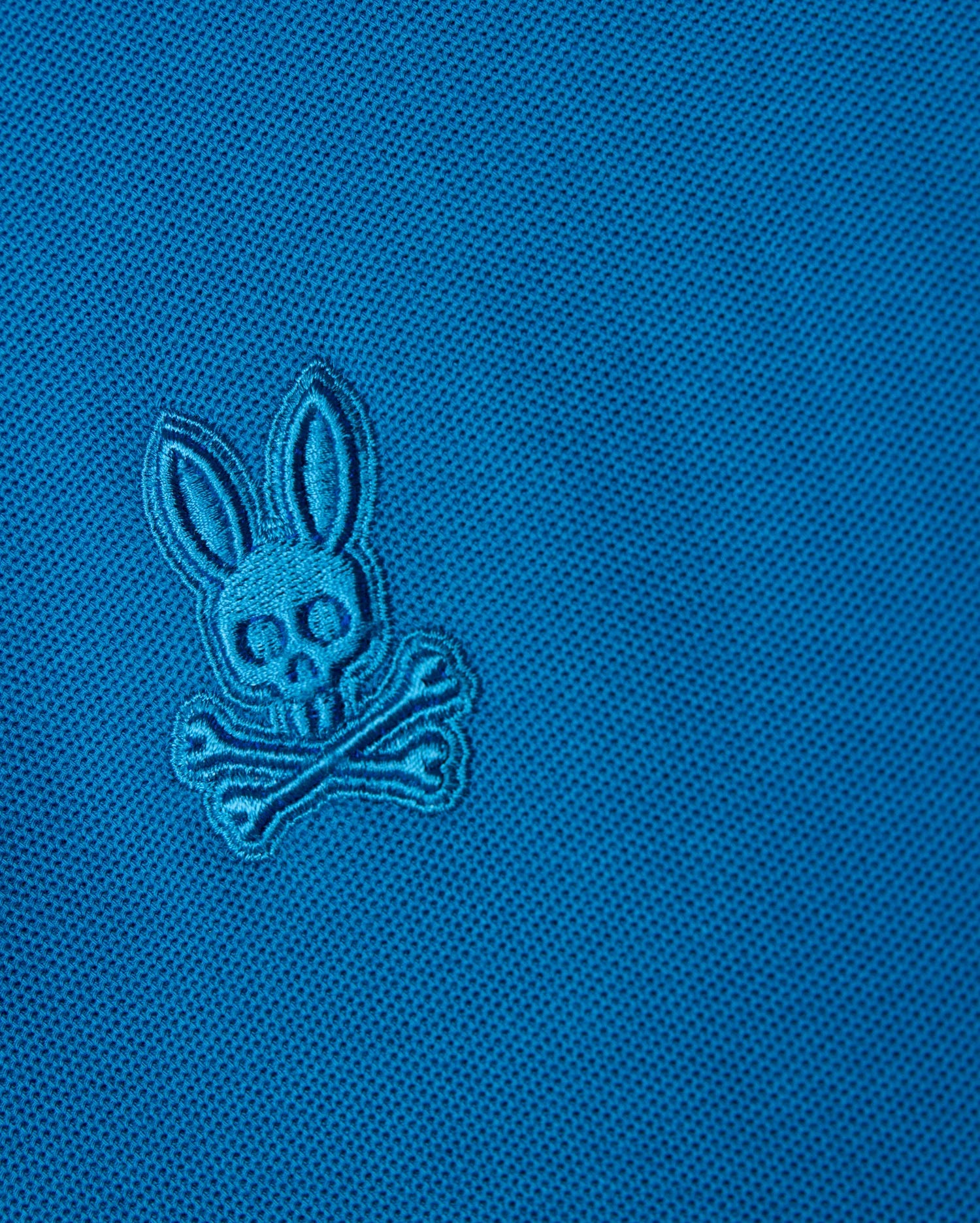 Close-up of the Psycho Bunny Kids Malton Pique Polo Shirt (B0K950D200) in luxurious Pima cotton piqué, featuring an embroidered logo with a stylized skull, bunny ears, and crossbones. The design is stitched in the same blue color as the fabric, creating a subtle, tonal effect. Mother-of-pearl buttons add an elegant finishing touch.