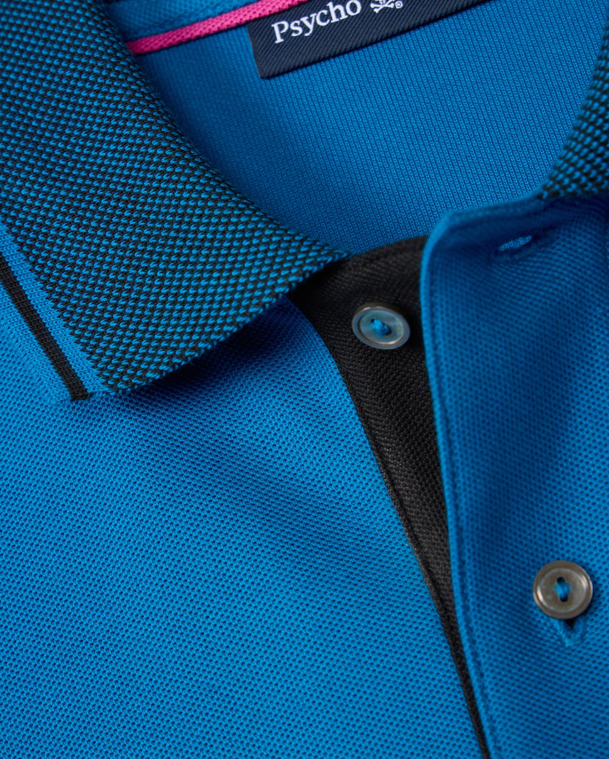 Close-up of a blue KIDS MALTON PIQUE POLO SHIRT - B0K950D200 with a textured collar featuring a black and white checkered pattern. The shirt has a button placket, and one mother-of-pearl button is visible. The brand label "Psycho Bunny" is seen on the inside of the collar, emphasizing high-quality Pima cotton piqué fabric.