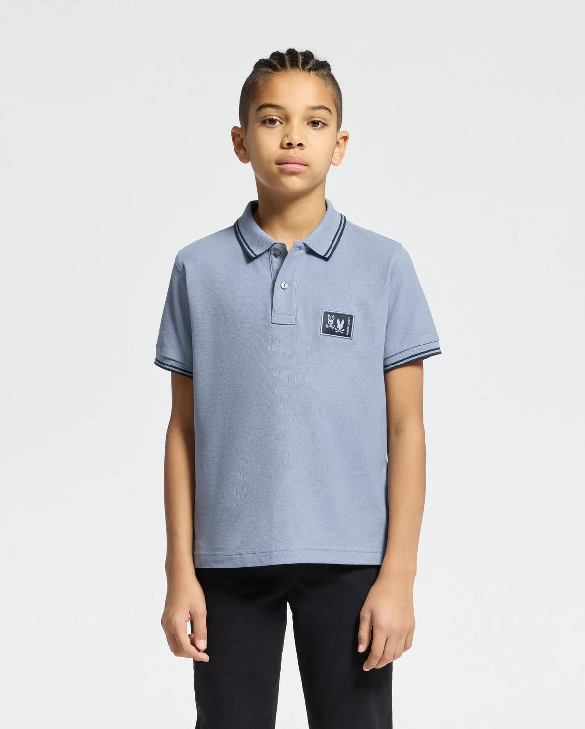 A young individual with braided hair wears the Psycho Bunny KIDS MONROE PIQUE POLO - B0K964D200, light blue in Pima cotton with a small logo appliqué, paired with black pants. The regular fit enhances their style against a plain white backdrop.