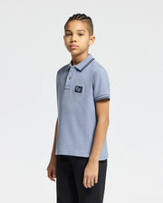 A young person with braided hair wears a Psycho Bunny KIDS MONROE PIQUE POLO in light blue, featuring a chest logo appliqué and regular fit. They're paired with black pants, standing relaxed against a plain white background.